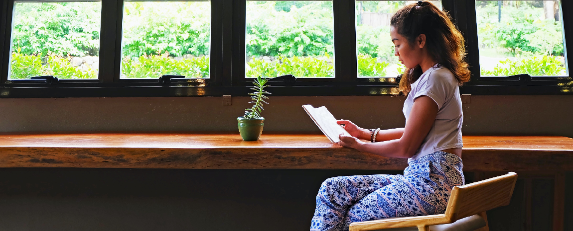 A woman sits at a wooden bar underneath windows reading.